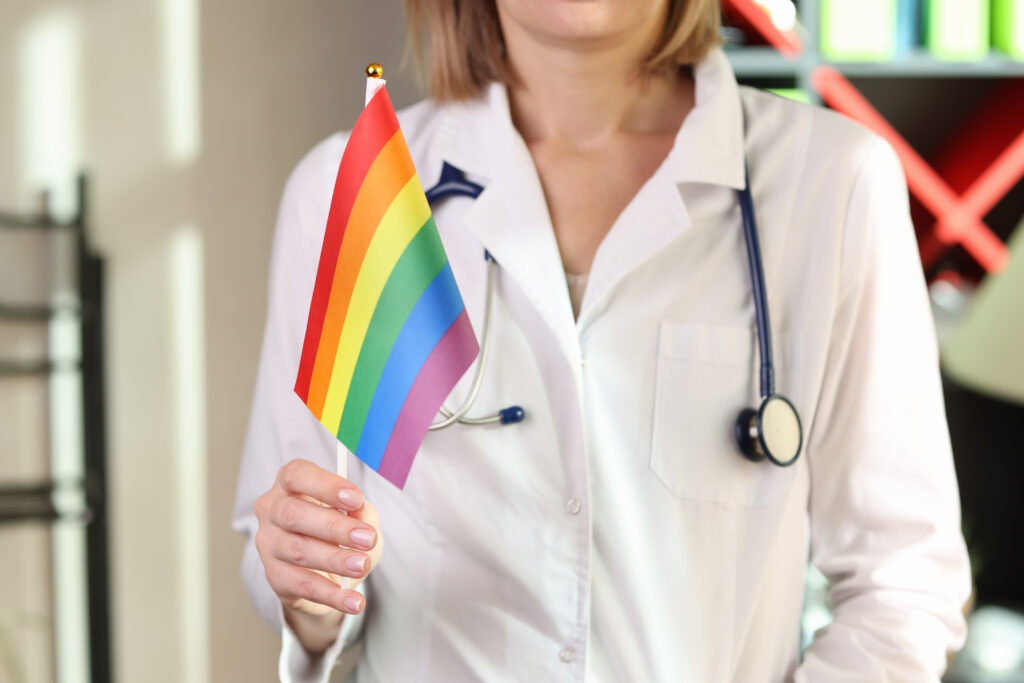 Profissional da saúde segura uma bandeira LGBTQIAP+, representando a dificuldade de acesso dessa população ao cuidado básico.